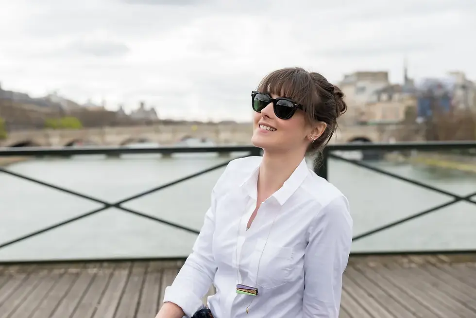 Photographe Chambéry - une femme souriante en chemise blanche et lunettes de soleil se tient sur un pont en bois. Paris en arrière-plan sous un ciel nuageux.