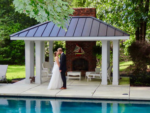 A couple kisses by a pool under a pavilion. They are dressed in wedding attire. A small cat stands nearby. Lush greenery surrounds them at Kateri Weddings and Events located in the Shenandoah Valley of Virginia