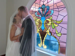 Bride and groom kiss under veil beside colorful stained glass window, set in a room with white walls, conveying romance and joy.