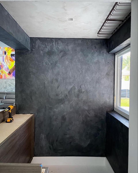 Interior wall with dark Venetian plaster finish in a modern space. The textured black plaster wall contrasts with a light ceiling and natural lighting from a nearby window. Contemporary home design with luxury wall finishes.