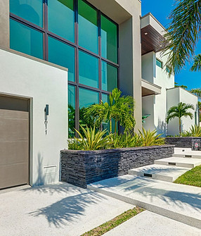 Modern luxury home with polished concrete walkway, lush landscaping, and sleek architectural design in a tropical setting.