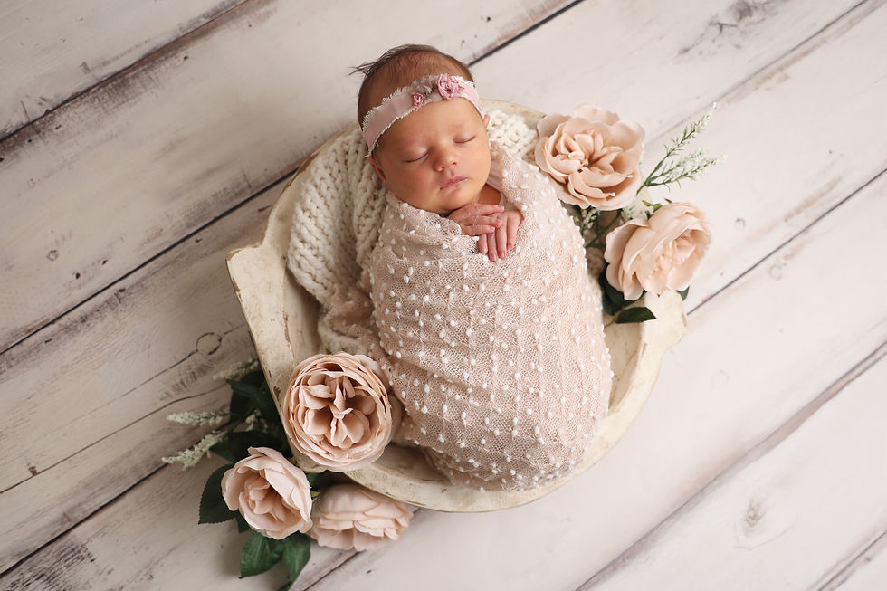 a newborn baby girl sleeping, wrapped in neutral colored wrap placed in a bowl with soft pink flowers and greenery surrounding it