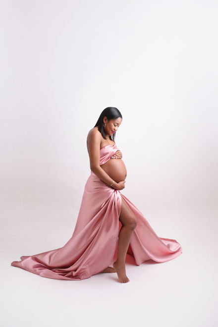 Pregnant woman in a flowing gown holding her belly, standing in soft light for a studio maternity portrait.