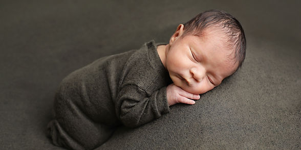 A baby boy in a dark green outfit sleeping on a dark green backdrop