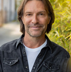 man in a casual button down shirt with white t-shirt standing outdoors. He las medium length blonde hair
