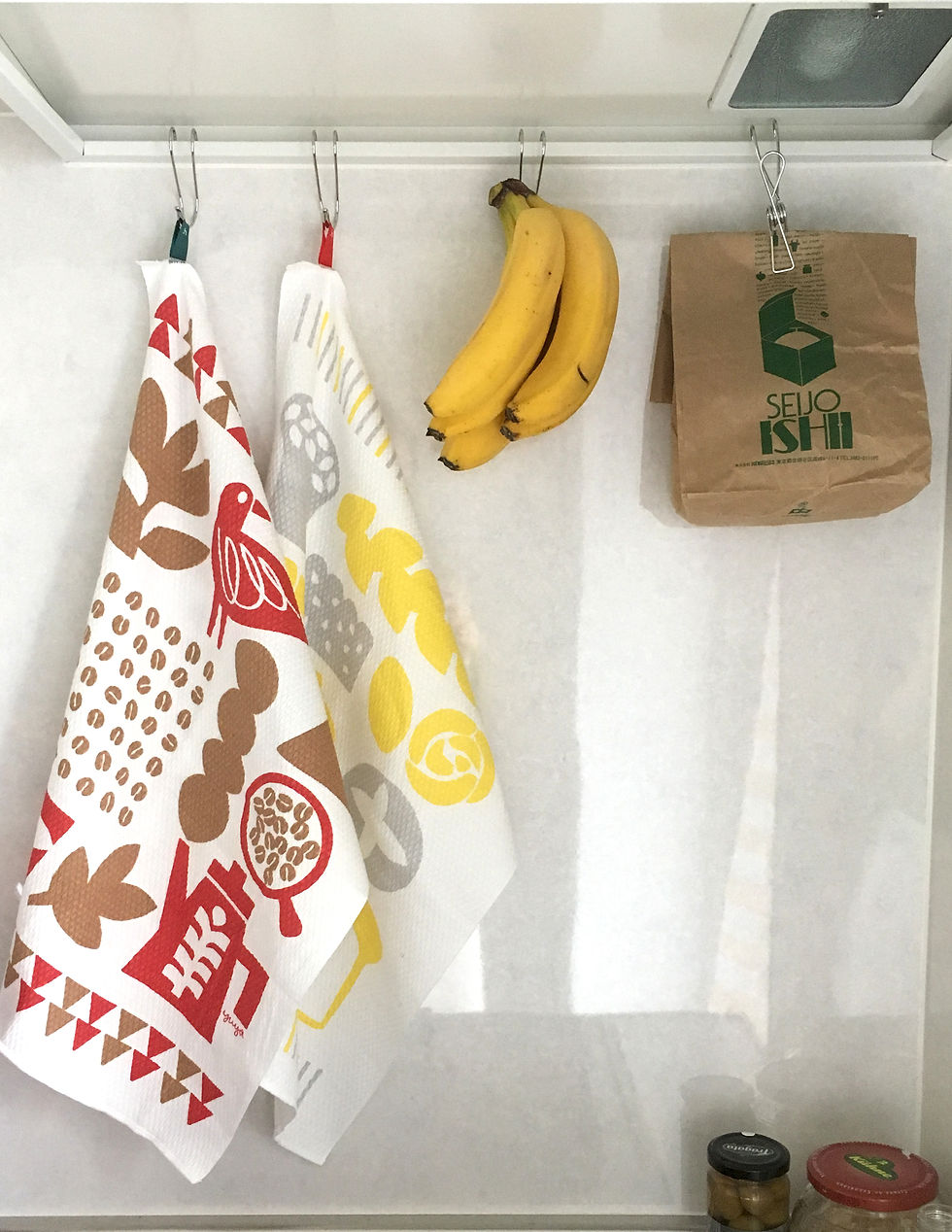 Thumbnail: Two Harada Orimono dishcloths — one with a pattern of coffee beans and a red coffee pot motif, the other with a yellow and grey pattern of bread motifs — both hanging on hooks alongside a bunch of bananas and a paper bag of coffee.