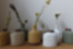 Eight mini vases designed by Angle in Japan all holding a dried flower. Each vase is a different in shape and colour, but all are flattened to create a relief like aesthetic. From left to right: mustard yellow, earthy stone, dusky blue-green, yellow, grey, two white, dusky green-blue.
