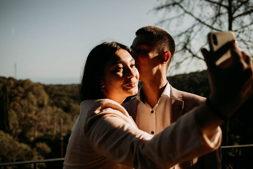 Primer plano de una pareja de invitados sonriendo mientras se toman un selfie con un teléfono móvil durante el atardecer en una boda en Girona; se aprecia un fondo de bosque y una iluminación cálida y lateral, por PICART.