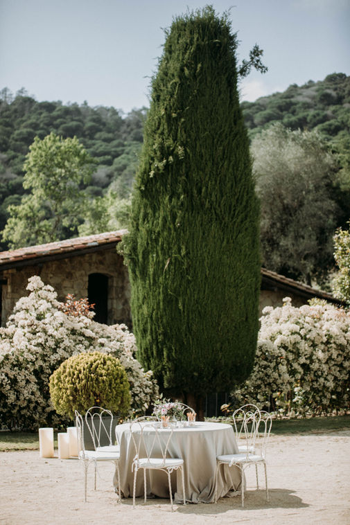 Mesa redonda con mantelería beige y sillas de hierro blanco dispuesta para un banquete de boda frente a un gran ciprés y una masía de piedra en Girona, bajo la luz natural del Empordán, por PICART.