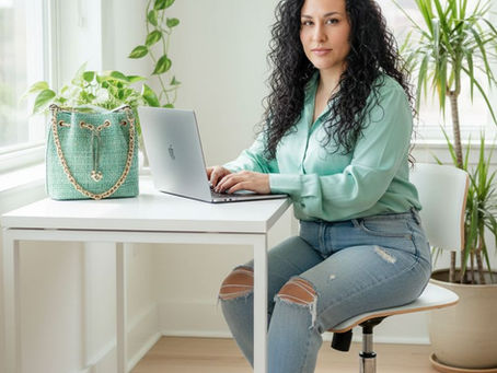 A woman with long, curly dark hair sits at a white desk in a bright, sunlit room, working on a silver laptop. She is wearing a light green button-down shirt, distressed blue jeans with ripped knees, and green snakeskin-patterned high heels. On the desk next to her is a textured light green handbag with a gold chain strap. The room is decorated with several lush green houseplants in the background, creating a fresh and professional atmosphere.