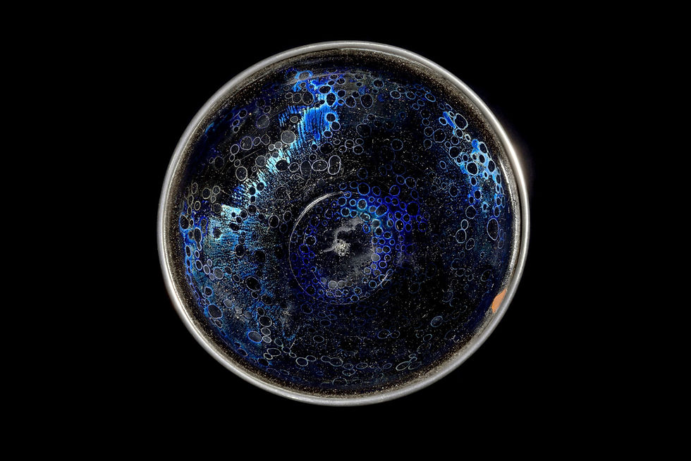 國寶南宋曜變天目茶碗 Top view of a deep blue ceramic bowl with iridescent bubble patterns on a black background, creating a cosmic, mysterious mood.