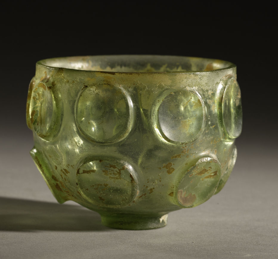 北周玻璃 Green glass cup with a circular pattern, slightly weathered. Set against a dark background, creating an ancient, rustic feel.