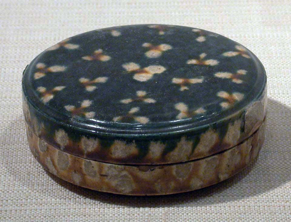 Round, lidded ceramic box with a dark, speckled pattern in beige on a textured surface. Set against a beige fabric background.