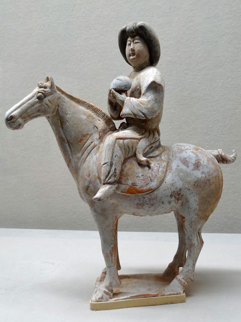 加彩騎馬婦人俑 Painted Pottery Figurine of Woman / A clay figure of a rider holding a ball sits on a horse. The weathered sculpture features earthy tones, and the rider wears a hat.