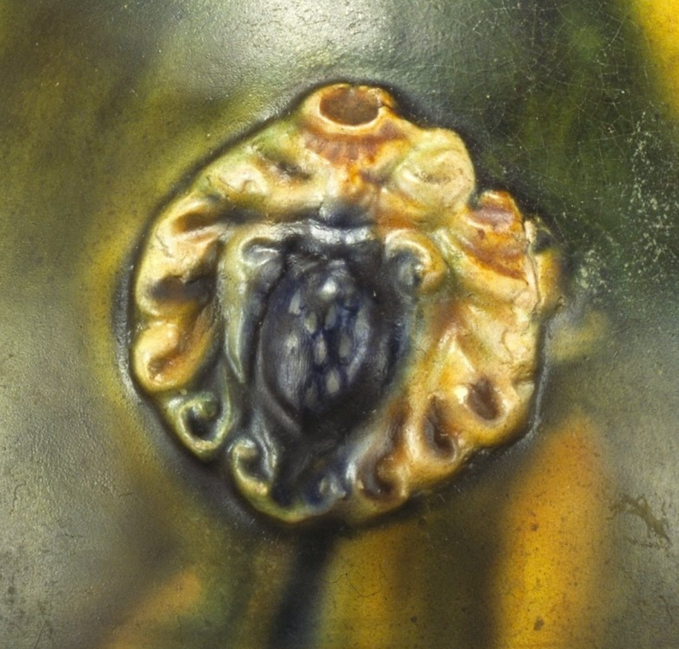 Close-up of a yellow and brown ceramic flower detail with a textured, cracked surface and green background, giving an aged look.