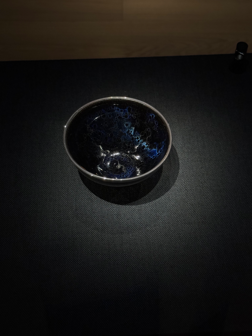 國寶南宋曜變天目茶碗 Ceramic bowl with intricate blue and black patterns sits on a dark textured table. Soft lighting highlights its glossy finish.