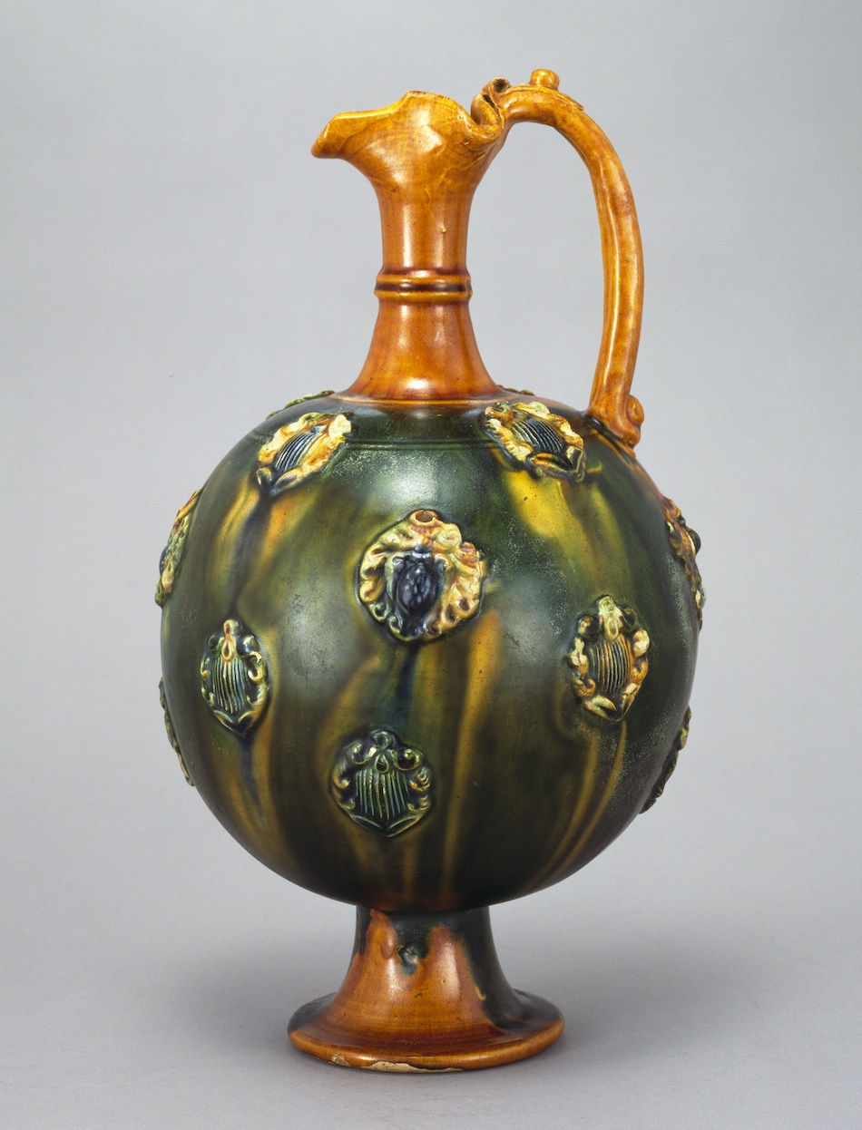 東京富士美術館:三彩貼花文水注 Ornate ceramic jug with a round body, decorated with intricate patterns. It features green and brown hues, set against a plain gray background.
