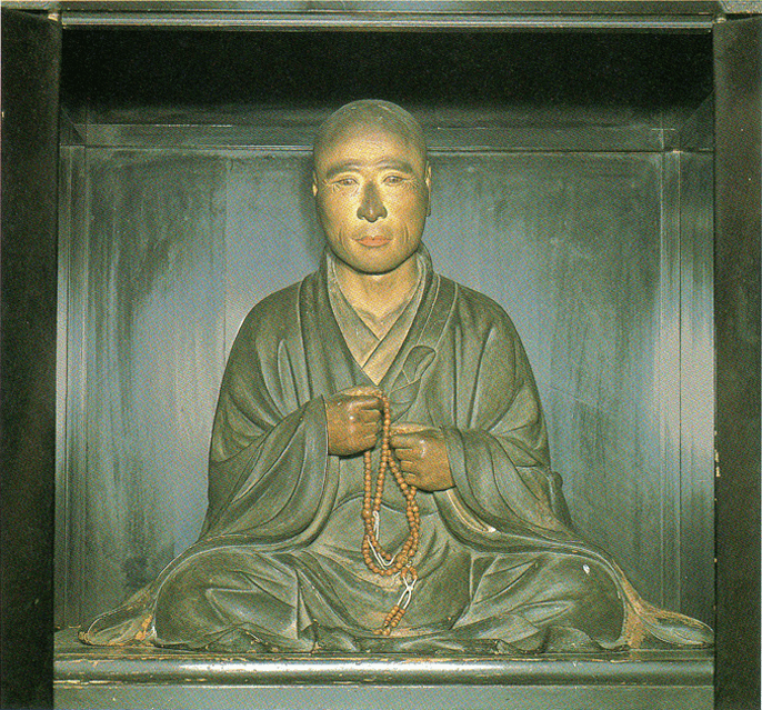 A wooden statue of a monk holding prayer beads, seated in meditation. The robe is dark, and the background is a shadowed, enclosed space.