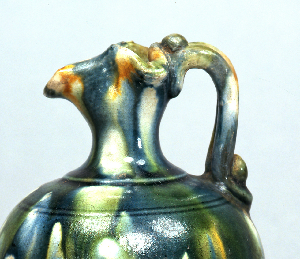 広田松繁,三彩藍釉水注 Close-up of a multicolored glazed ceramic jug with a distinct handle against a plain background. The colors include blue, green, and orange.