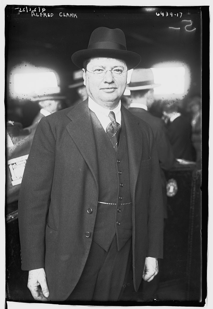 Man in a suit and bowler hat stands in a busy indoor setting. Text above reads "Alfred Clark." Black and white image, early 20th-century style.