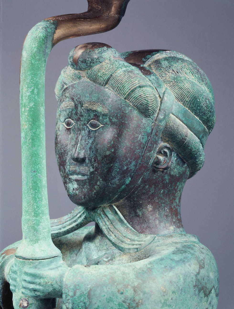 Close-up of a weathered bronze statue with green patina, holding a staff against a plain gray background.
