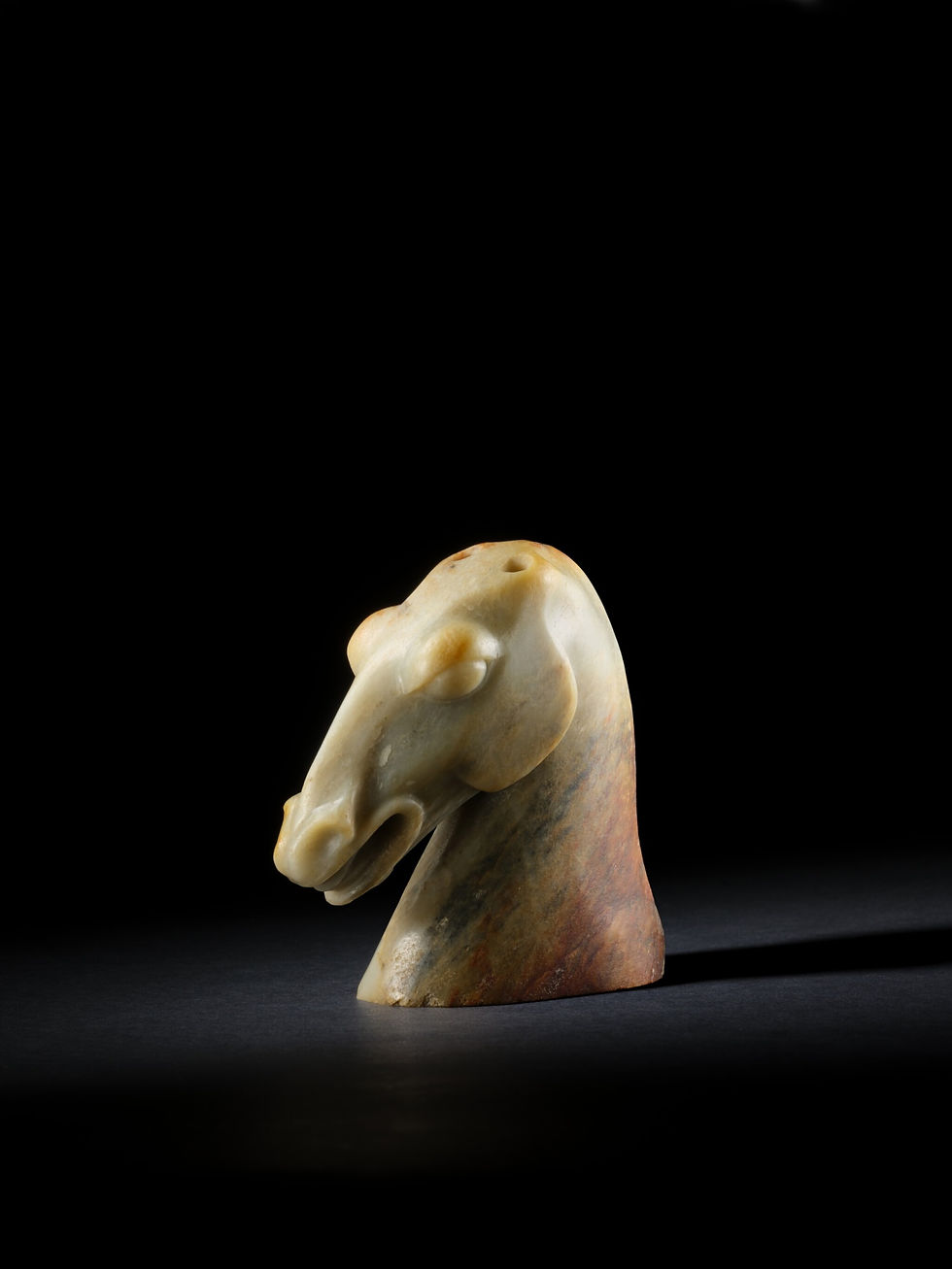 Carved stone horse head sculpture in beige and brown, isolated on a black background with dramatic lighting.