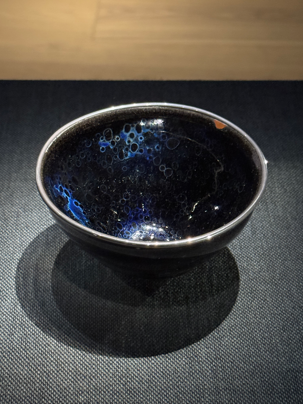 國寶南宋曜變天目茶碗 A dark ceramic bowl with blue bubble patterns sits on a textured surface under soft lighting, creating a calm, artistic ambiance.