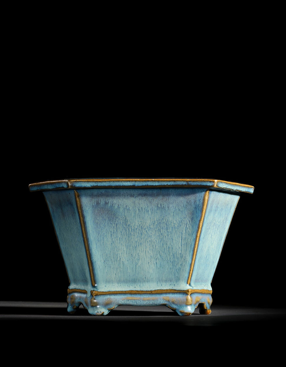 Blue ceramic pot with a rectangular shape and ornate base on a black background. Subtle gold accents highlight edges. Minimalist style.