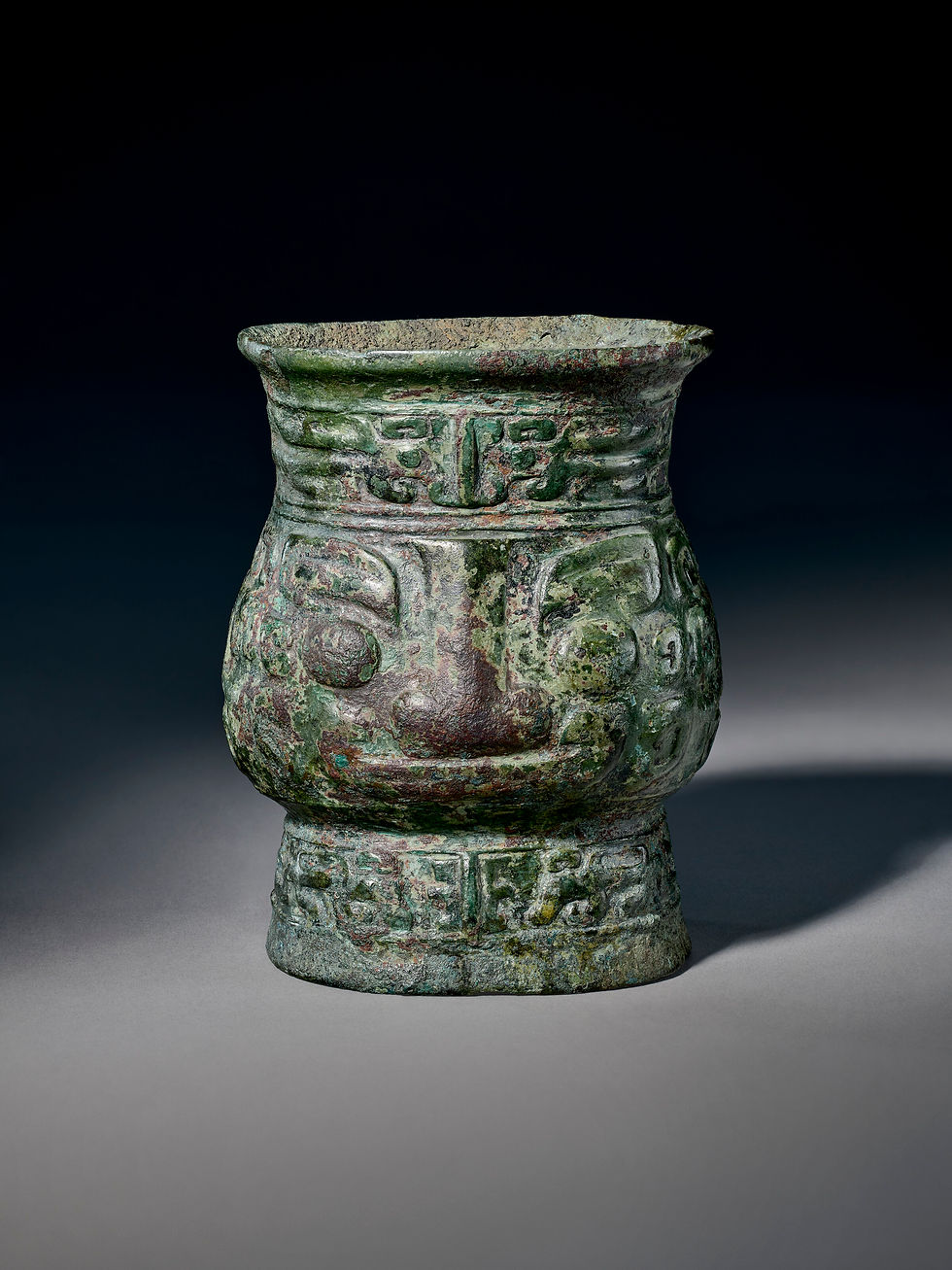 Ancient green bronze vessel with intricate carvings, resembling a smiling face, against a dark background.
