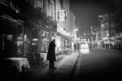 Man stands outside restaurant, clock above entrance, outdoor seating, city street at night. Image (c) Lenny Di Lorenzo.