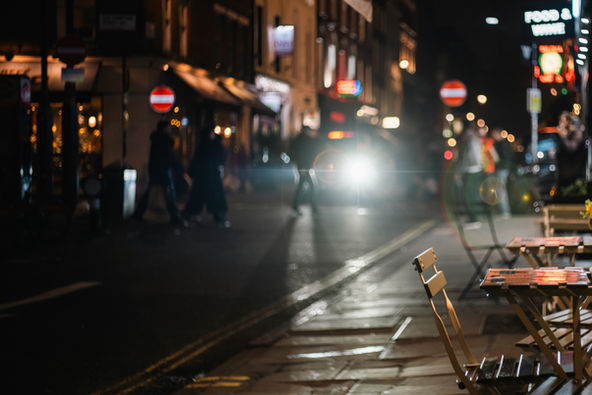 A city street at night outside a cafe, a chair in the foreground, car lights and people walking in the background.   Image (c) Lenny Di Lorenzo.