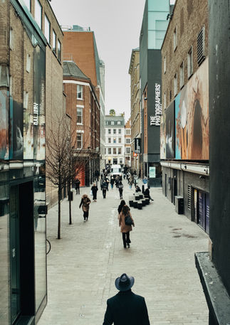 People walk in a London street, a man wearing a hat is visible in the foreground as he ventures into it. Image (c) Lenny Di Lorenzo.
