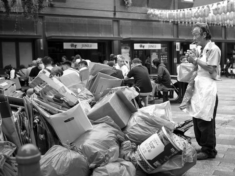 A Chinese man lights up a cigarette in front of a big pile of refuse in Chinatown, London. Image (c) Lenny Di Lorenzo.