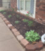 Flower bed with blooming red roses and green plants on mulch, bordered by bricks. Located next to a brick house with blue shutters.