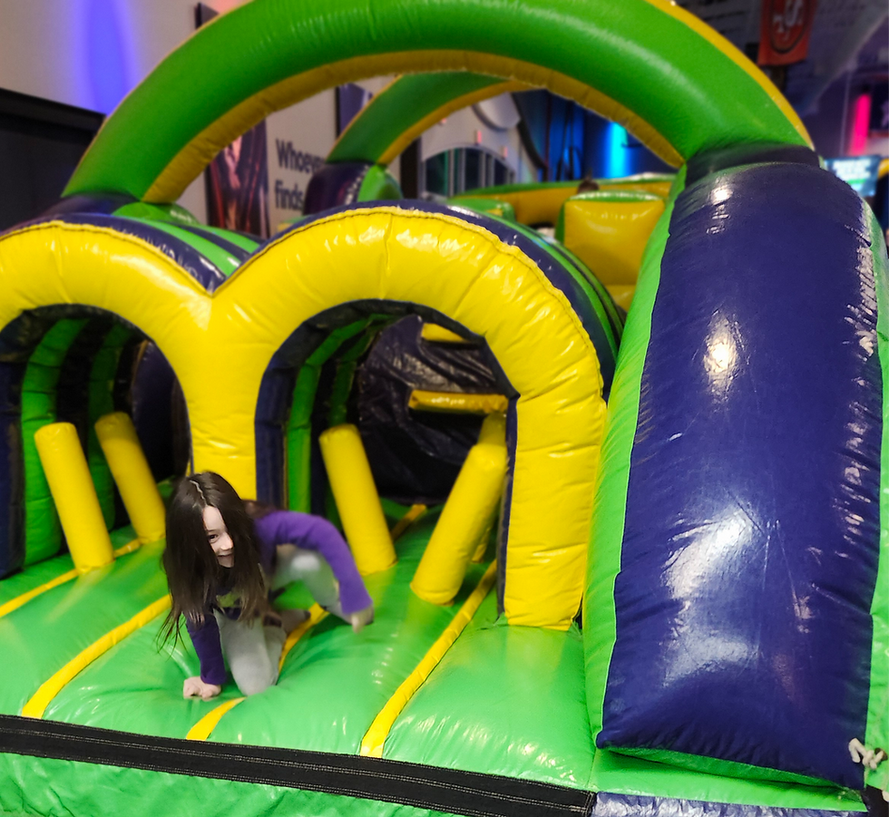 Child crawls through a green and yellow inflatable obstacle course indoors. A hand waves nearby. Bright, playful atmosphere.