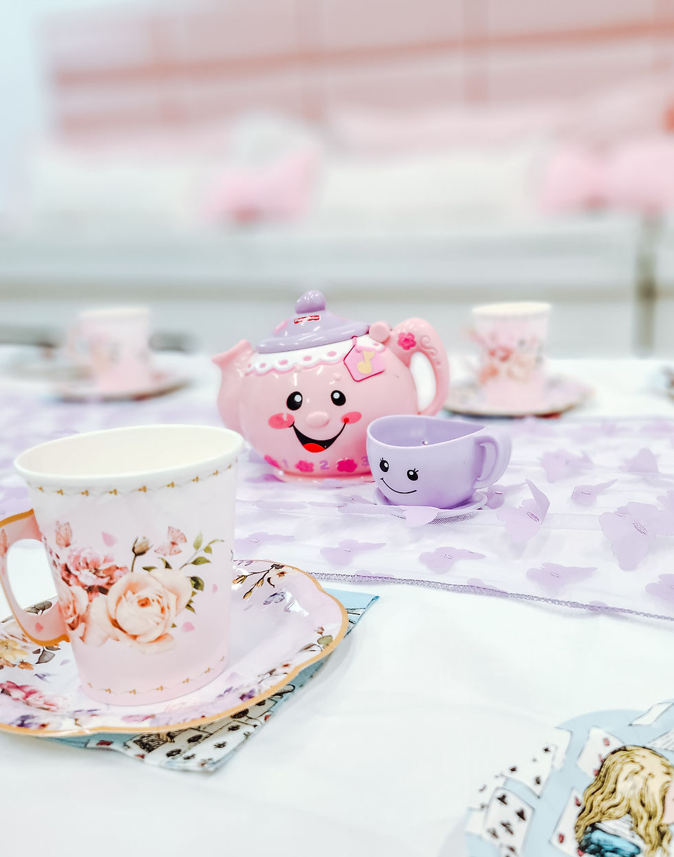 Cute pink teapot and cups with smiling faces on a pastel tablecloth. Floral pattern cups and saucers create a playful, cheerful vibe.