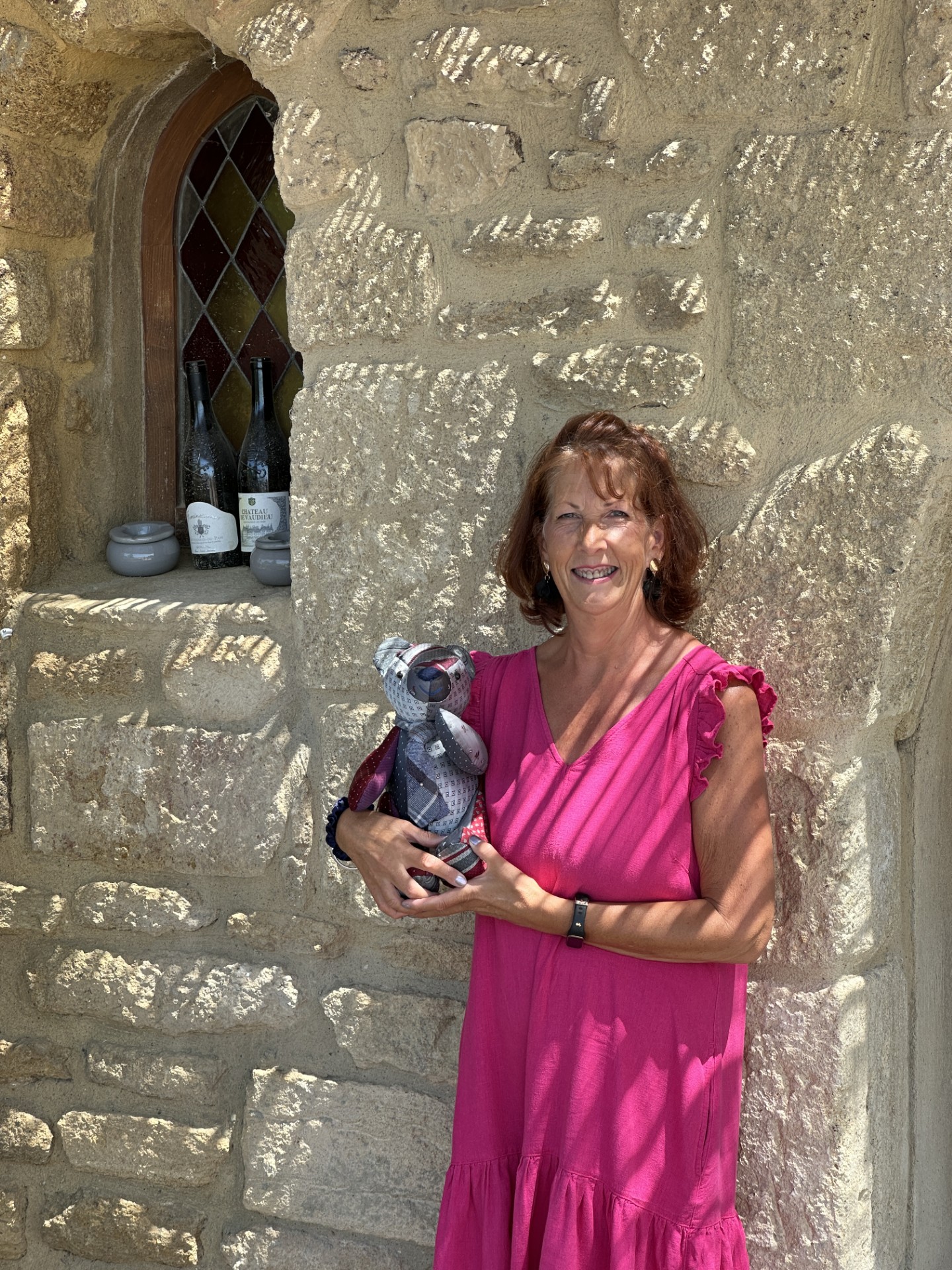 Sandi holding Gordon Bear in Les Caves Saint Charles, standing by rustic stone walls and wine bottles, embodying the warmth of Châteauneuf’s wine journey.