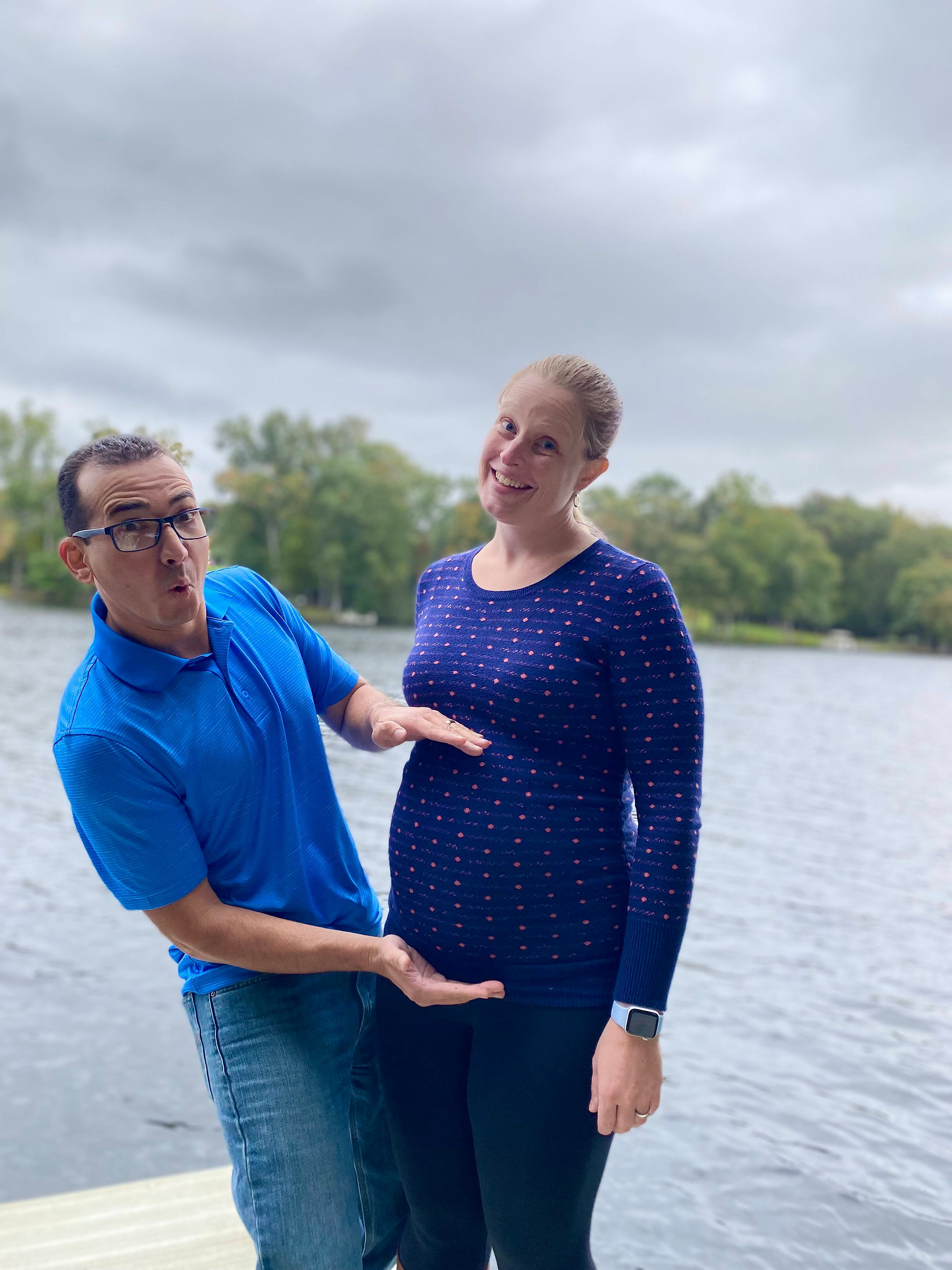 Husband and wife pose for maternity photo. Husband makes goofy face and frames the pregnant belly with his hands, wife laughs