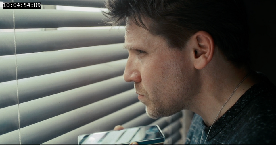 Eine dramatische Nahaufnahme im Profil von einem Mann in einer Krimi-Produktion, der angespannt durch die Lamellen einer grauen Jalousie nach draußen späht. Er hält dabei ein Smartphone nah an sein Gesicht.