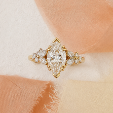 Styled view of the Calanthe yellow gold engagement ring, featuring a marquise-cut lab-grown diamond and delicate floral diamond shoulders on blush and ivory silk.