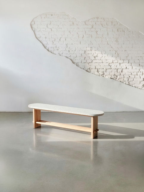 Trini Bench with a terrazzo seat on a pale wood frame in front of a white brick wall