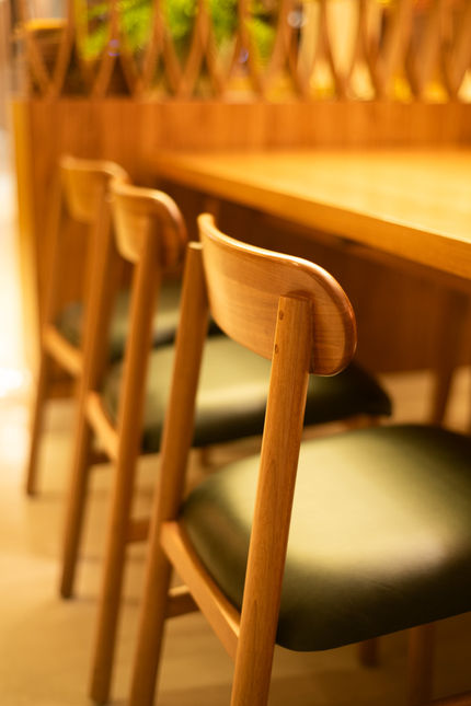 Row of Hi Chairs in light solid wood with dark green upholstered seats along a long wooden table in a warmly lit restaurant