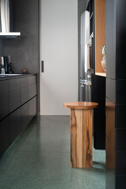 Trini Bar Stool in warm wood standing alone in a dark kitchen with terrazzo flooring