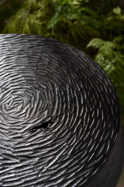 Macro of a blackened Ybyrá Stool seat, concentric adze marks radiating from a natural knot, with foliage behind
