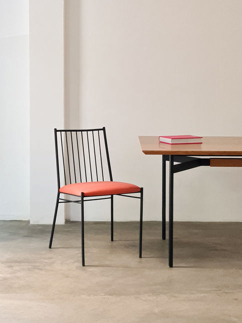 Joaquim Chair with a coral seat beside a wooden table holding books, on a polished concrete floor
