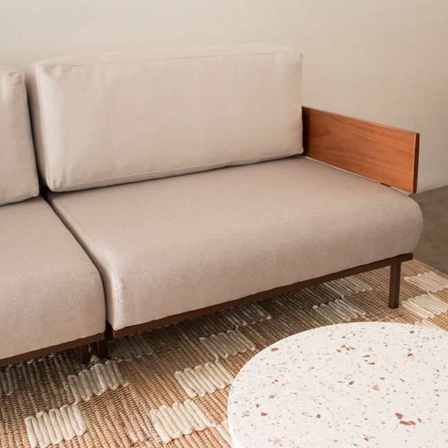 Indy Sofa in soft beige upholstery with a solid wood arm, on a textured rug next to a round terrazzo coffee table
