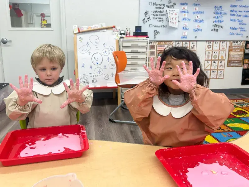 Children with pink hands from a craft activity sit at a table with trays of paint. Classroom setting with drawings and text on the wall.