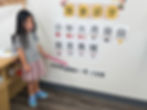 Young girl in a gray shirt points at Chinese characters on a classroom wall. She's wearing a pink skirt and colorful shoes, standing by a table.