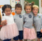 Four smiling children in a classroom wearing gray shirts and pink tutus. They stand close together against a colorful decor background.