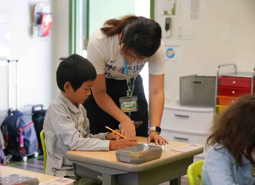 Teacher assists student writing at a desk in a colorful classroom. Calm atmosphere with school supplies in the background.